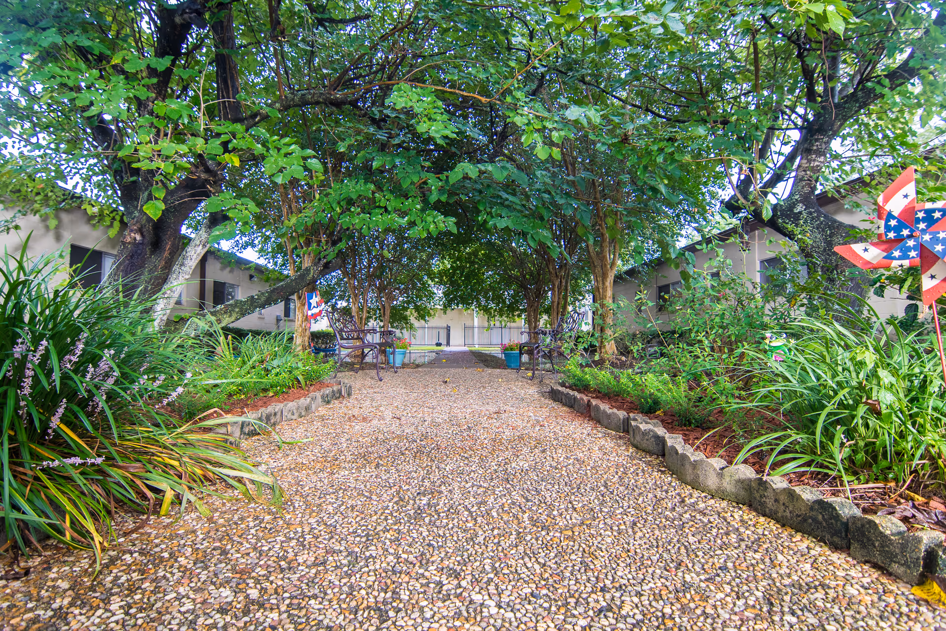A gravel pathway lined with plants and flowers on both sides, leading to a shaded area with benches under large leafy trees. There are buildings visible on either side of the pathway and a decorative pinwheel on the right side.