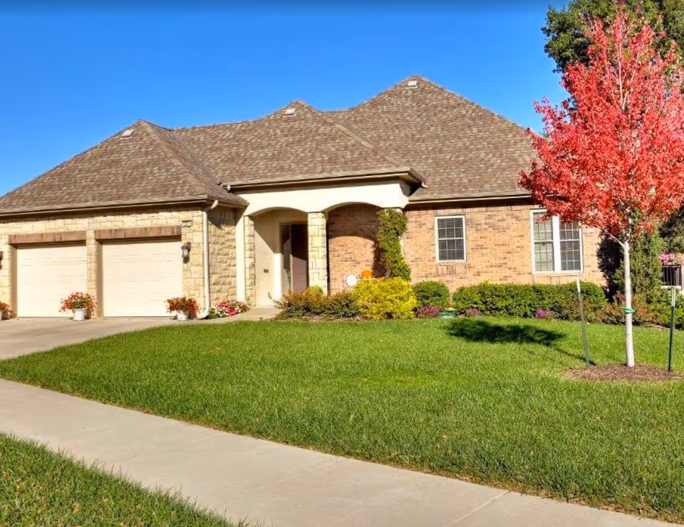 Exterior view of a single-story brick and stone house with a two-car garage, a well-maintained green lawn, colorful flower pots near the garage, and a vibrant red-leaved tree on the right side under a clear blue sky.