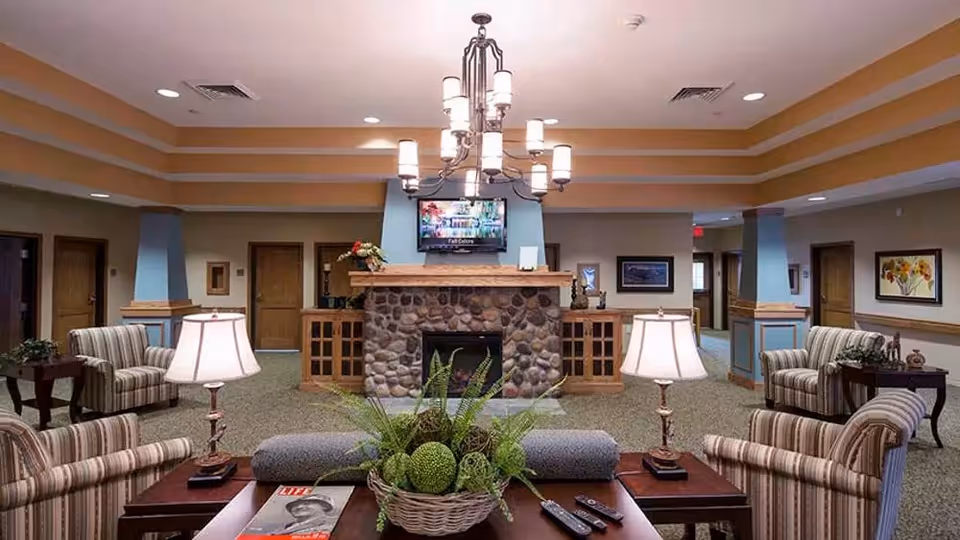 A cozy living room area in a senior living facility featuring a stone fireplace with a TV mounted above it. The room has striped armchairs arranged around a wooden coffee table with a decorative plant and magazines. Two table lamps provide warm lighting, and the walls are decorated with framed artwork. The ceiling has recessed lighting and a chandelier.