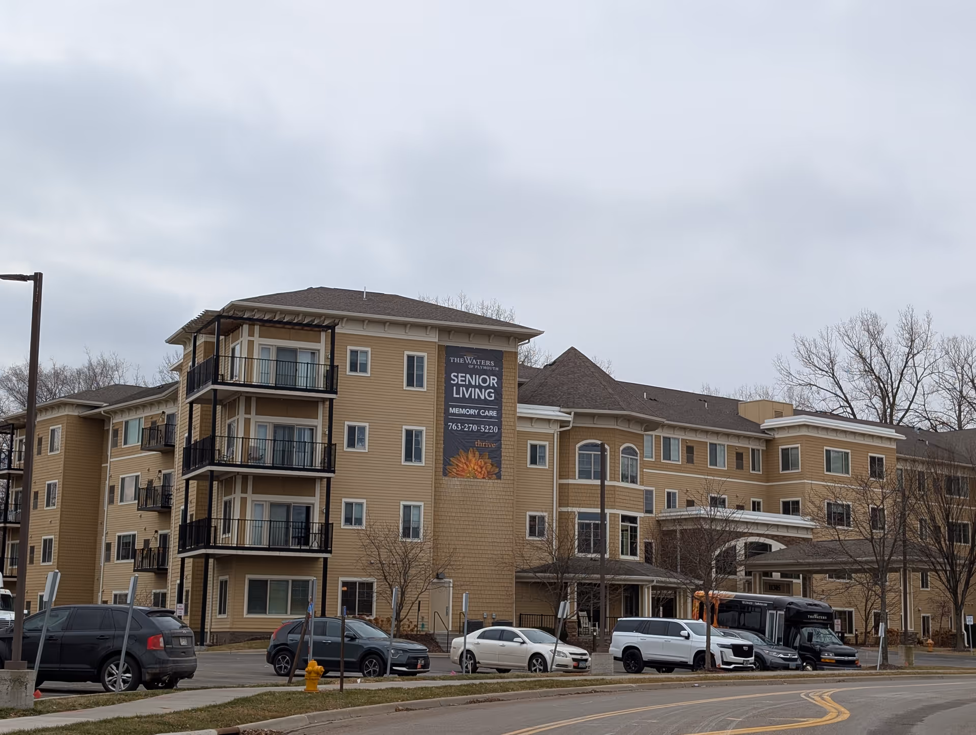 Exterior view of a multi-story senior living facility named The Waters of Plymouth with several parked cars in front and leafless trees around the building under a cloudy sky.