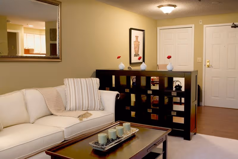 A cozy living room area with a white couch adorned with a striped pillow and a beige throw blanket. In front of the couch is a wooden coffee table with a decorative tray holding five candles. Behind the couch is a dark wooden room divider with square cutouts, decorated with three small white vases each holding a single red flower. A framed picture hangs on the beige wall above the divider. The room has a hardwood floor near the entrance door and a white carpet in the living area. A large mirror reflects part of a kitchen area in the background.