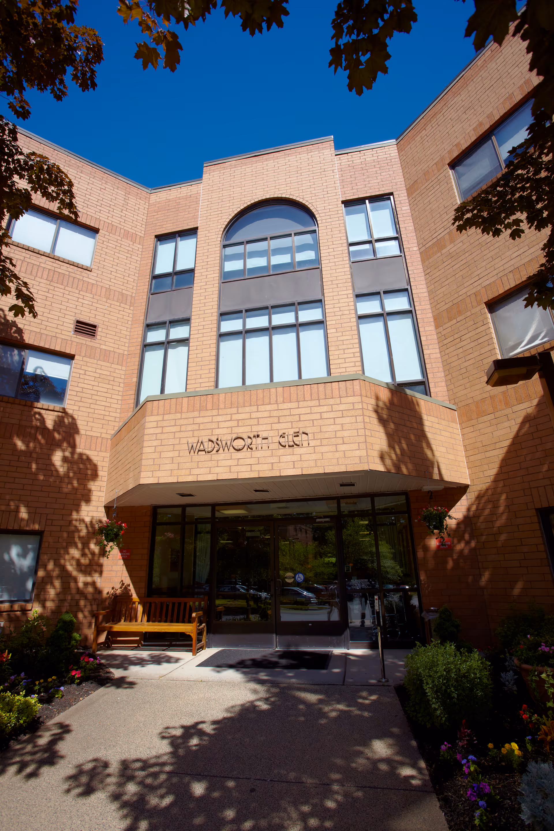 Exterior view of the entrance to Wadsworth Glen Health Care and Rehabilitation Center, a brick building with large windows and a glass door entrance. There are trees casting shadows and some flowers and greenery near the entrance, along with a wooden bench.