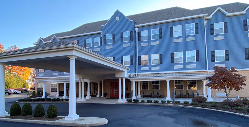 Covered porte-cochère and front façade of a three-story blue assisted living building with white columns and a circular drive.