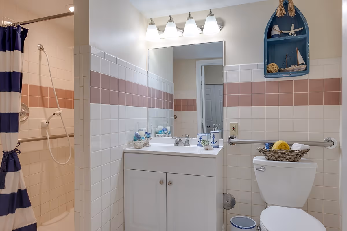 A bathroom with white and pink tiled walls, featuring a white vanity with a sink and a large mirror above it. There is a toilet with a basket containing a yellow sponge and towels on top. A blue nautical-themed wall shelf holds decorative items. The shower area has a blue and white striped curtain and a handheld showerhead. A grab bar is installed next to the toilet.