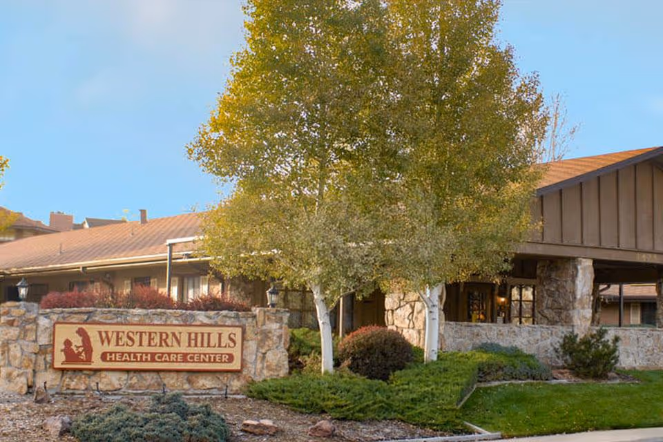 Front entrance of Western Hills Health Care Center showing a stone sign, landscaped shrubs, and trees in front of the building.