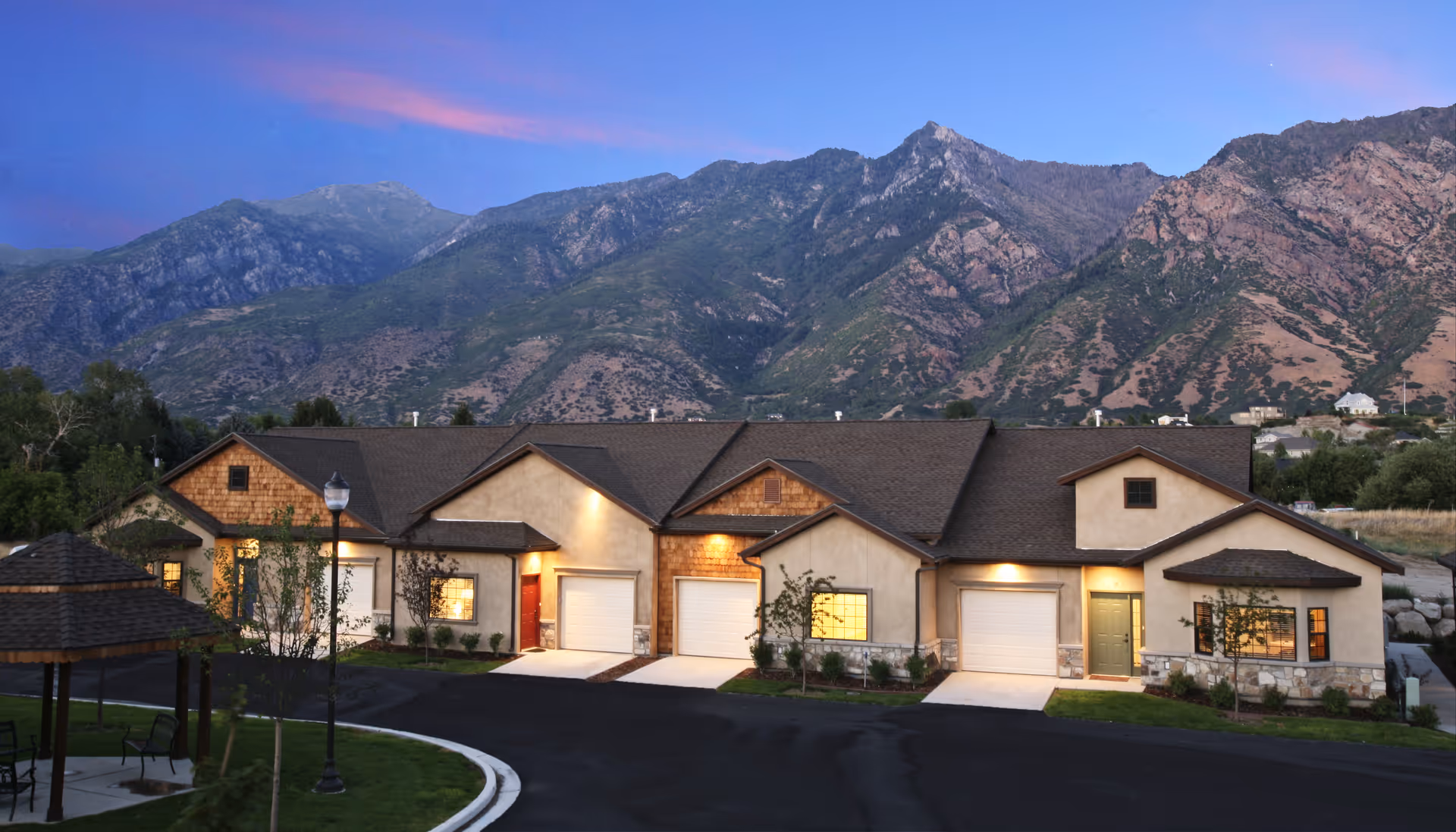 Row of attached single-story residences with lit garages and a gazebo in front, set against a mountain backdrop at dusk.