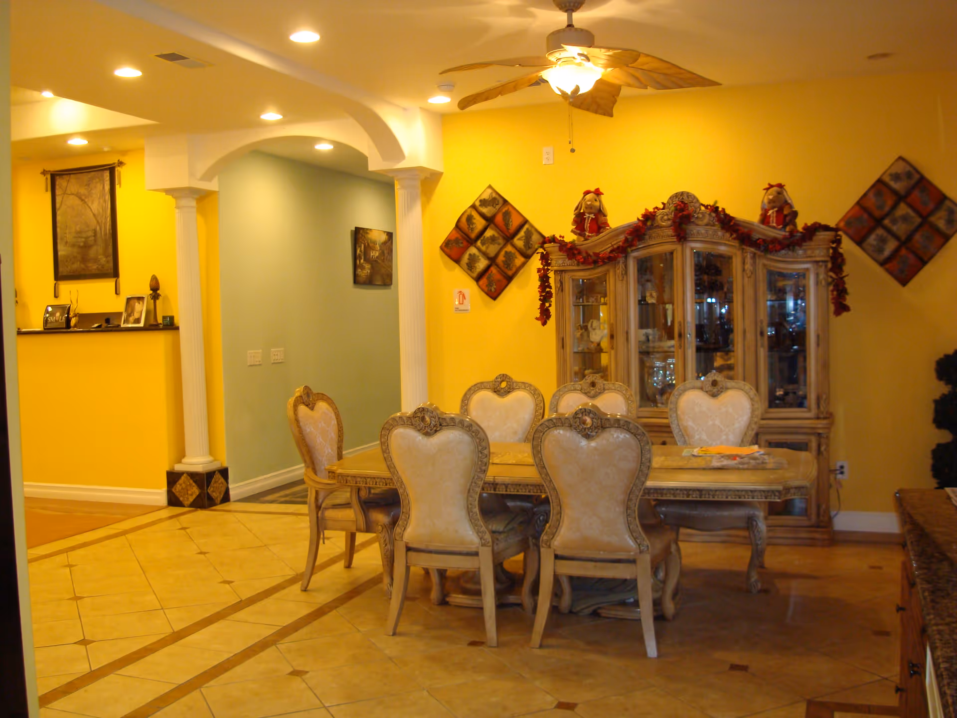 A dining room with a large wooden dining table surrounded by eight ornate upholstered chairs. Behind the table is a glass-fronted china cabinet decorated with garlands and two doll figures on top. The walls are painted yellow and light blue, with decorative wall hangings. The floor is tiled, and a ceiling fan with a light fixture is above the table.