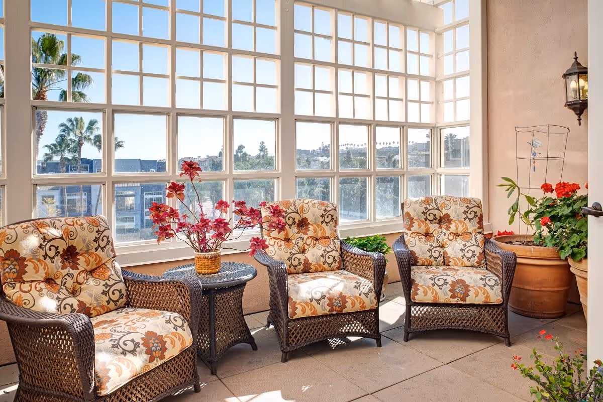 A sunlit indoor seating area with three cushioned wicker armchairs arranged around a small wicker table holding a vase with red flowers. Large grid-style windows provide a view of palm trees and buildings outside. There are also potted plants placed around the seating area.