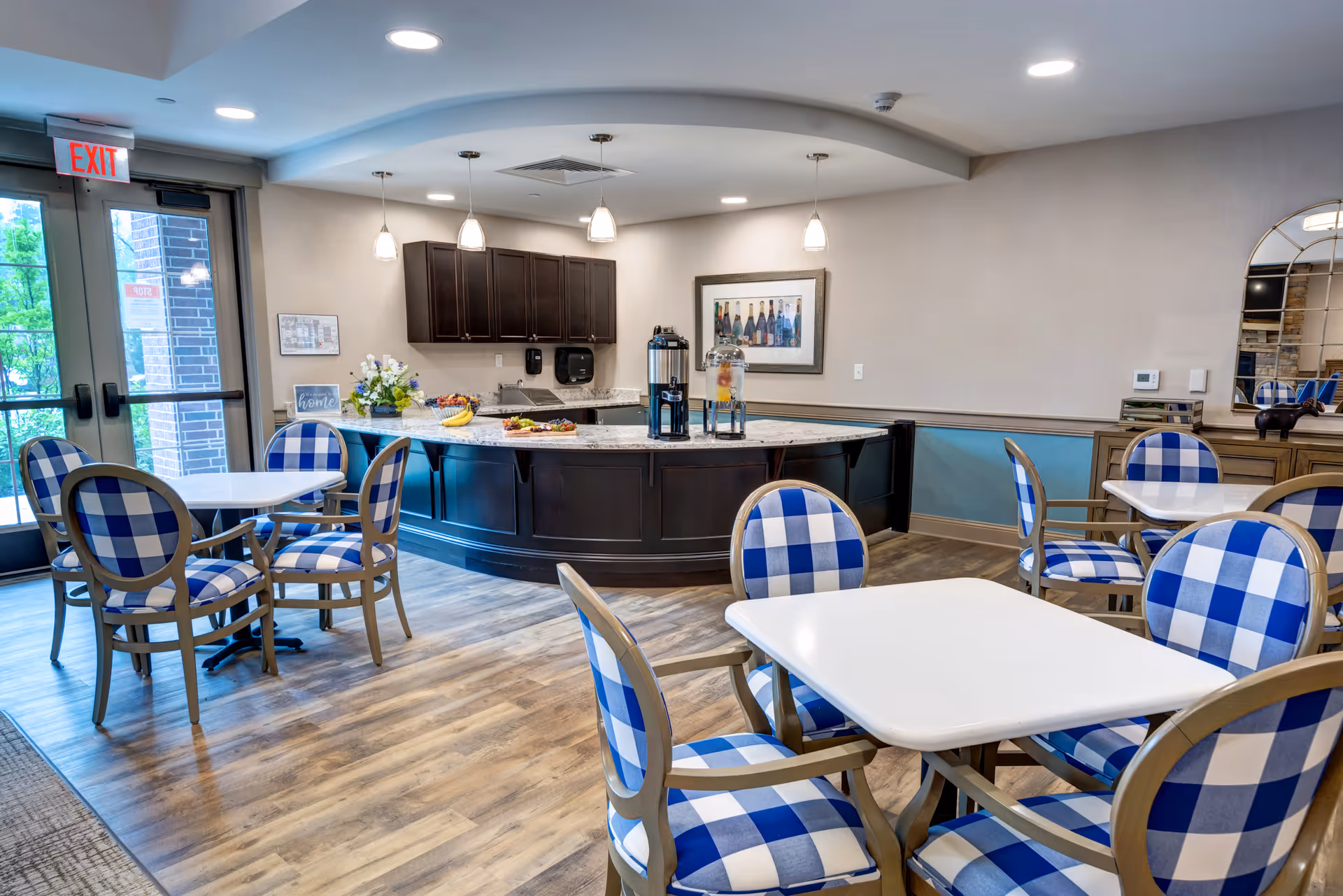 A bright and inviting dining area in a senior living facility featuring several white tables surrounded by chairs with blue and white checkered cushions. In the background, there is a curved counter with dark wood cabinetry, a coffee dispenser, and a water dispenser. The room has wood flooring, light-colored walls, and a large windowed door with an exit sign above it.