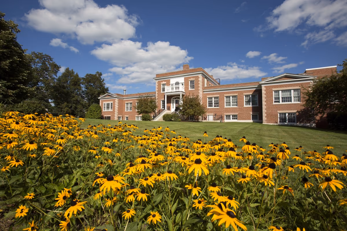 A large brick building with white trim under a blue sky with scattered clouds. In the foreground, there is a lush green lawn with a dense patch of yellow flowers. Trees surround the building on both sides.