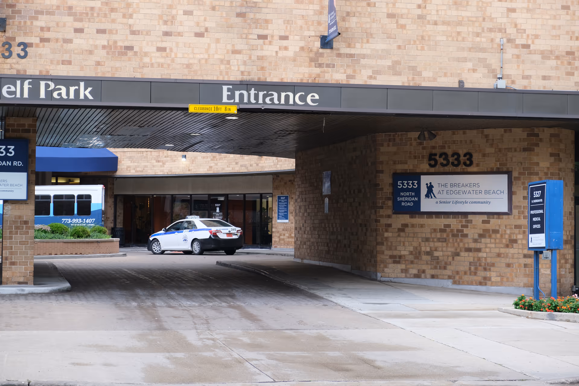 Entrance driveway of The Breakers at Edgewater Beach senior living community, showing a covered drop-off area with a taxi parked near the entrance. The building is made of light brown bricks and has signs displaying the address 5333 North Sheridan Road and the facility name.