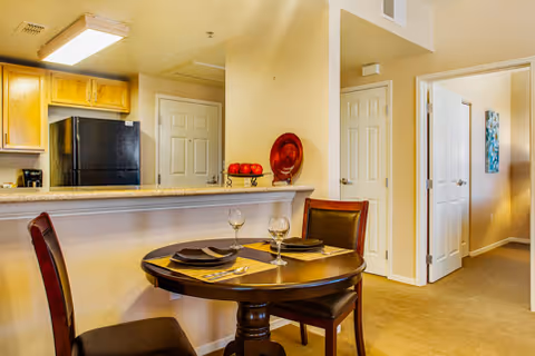 A cozy dining area with a round wooden table set for two with placemats, plates, and wine glasses. Behind the table is a kitchen with wooden cabinets, a black refrigerator, and a coffee maker. The walls are painted beige, and there are two white doors visible in the background along a hallway.
