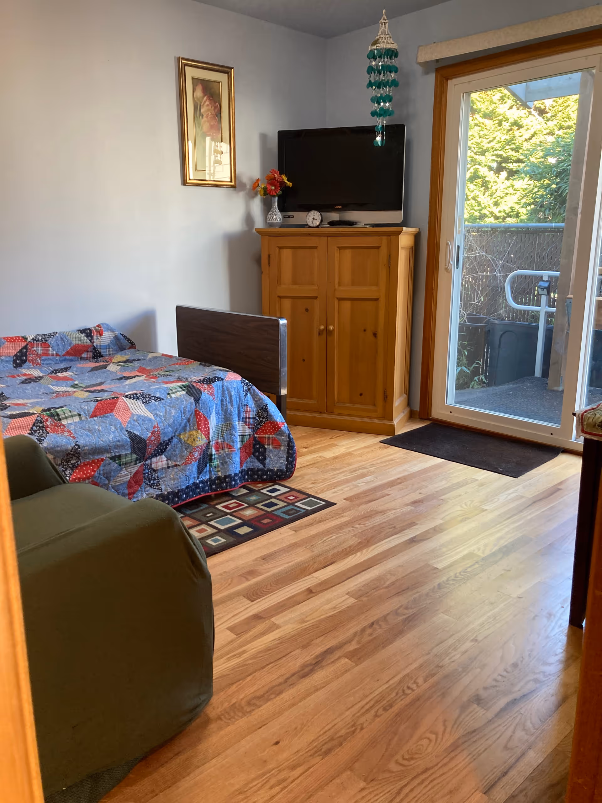 A cozy bedroom with a colorful patchwork quilt on the bed, a green armchair, a wooden cabinet with a TV on top, a small clock and a vase with flowers. There is a framed picture on the wall and a sliding glass door leading to an outdoor area with greenery visible outside.