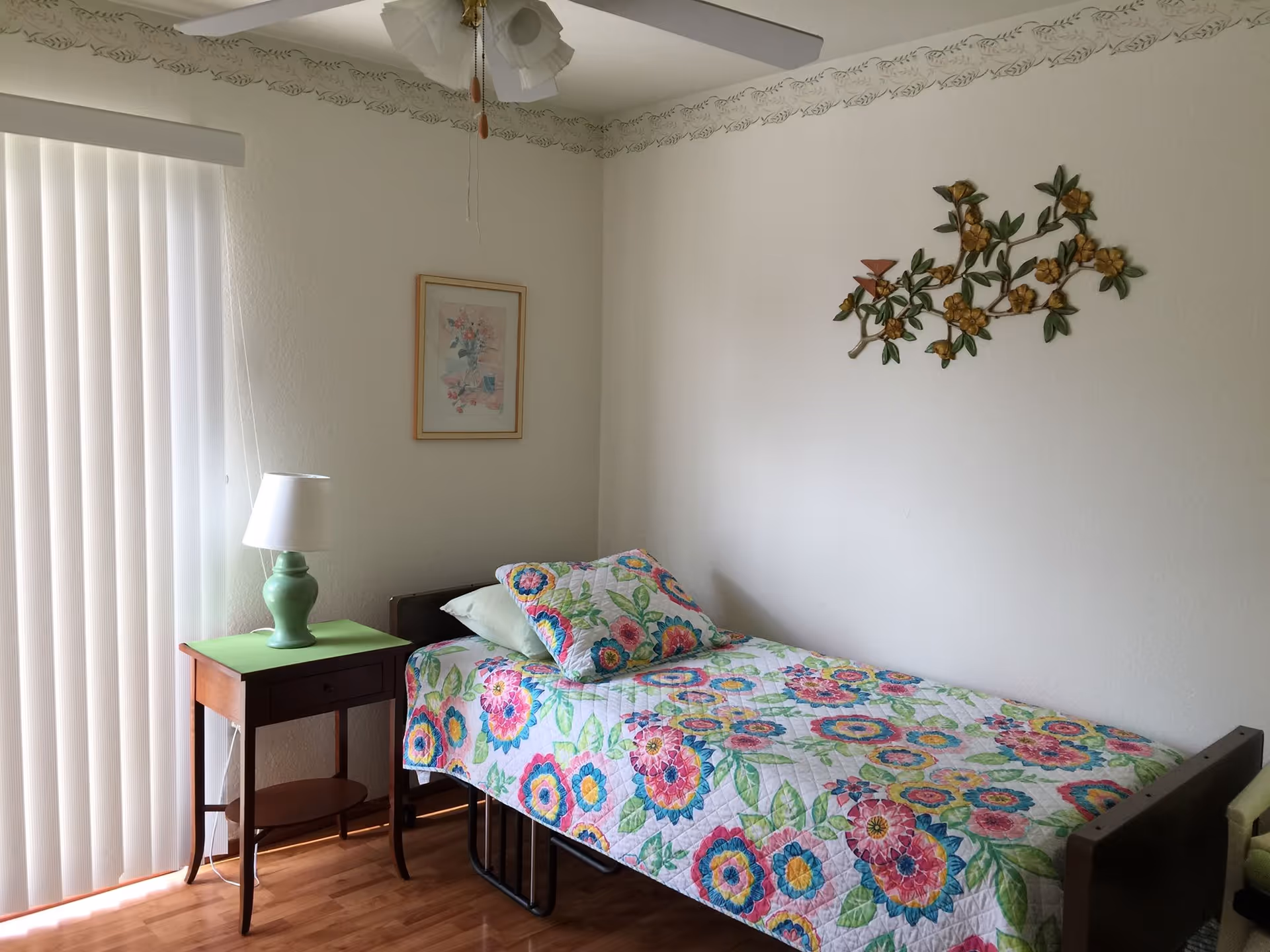 A small bedroom with a single bed covered in a colorful floral quilt and matching pillow. Next to the bed is a wooden nightstand with a green table lamp. The room has light-colored walls with a floral metal wall decoration and a framed picture. Vertical blinds cover a window or sliding door, and a ceiling fan is visible above.