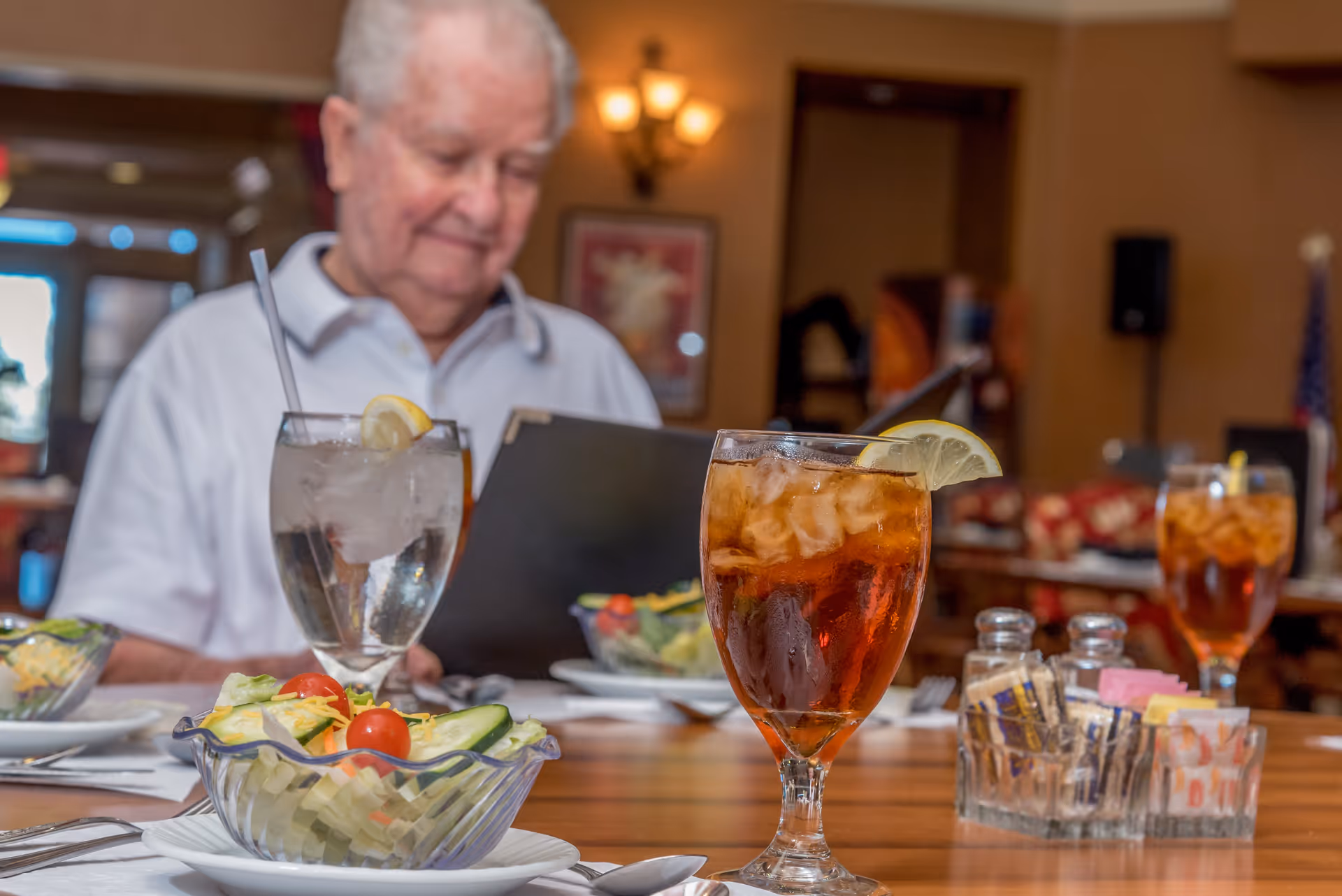 An elderly man sitting at a dining table in a restaurant or dining room, looking at a menu. On the table in front of him are glasses of iced tea with lemon, a glass of water with lemon, and bowls of salad with cherry tomatoes and cucumber slices. The background shows warm lighting and framed artwork on the wall.