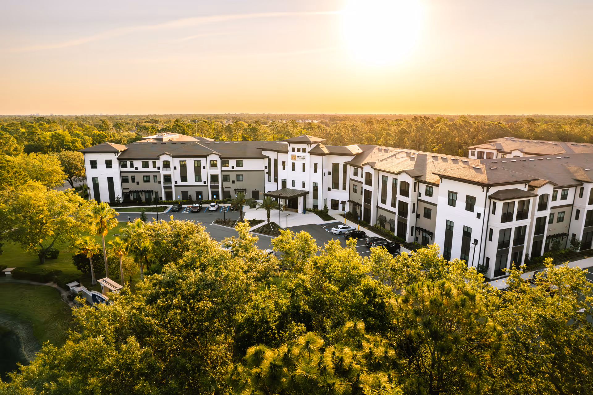 Aerial view of a large, modern senior living facility building surrounded by lush green trees under a bright, setting sun sky.