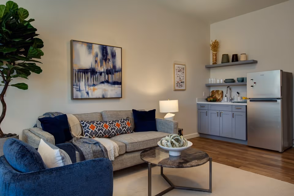 A cozy living area featuring a gray sofa with patterned and solid pillows, a blue armchair, a round coffee table with decorative items, a floor lamp, and a kitchenette with gray cabinets, a sink, open shelves with dishes, and a stainless steel refrigerator. A large abstract painting and a smaller framed artwork hang on the beige walls, and a tall green plant is in the corner.