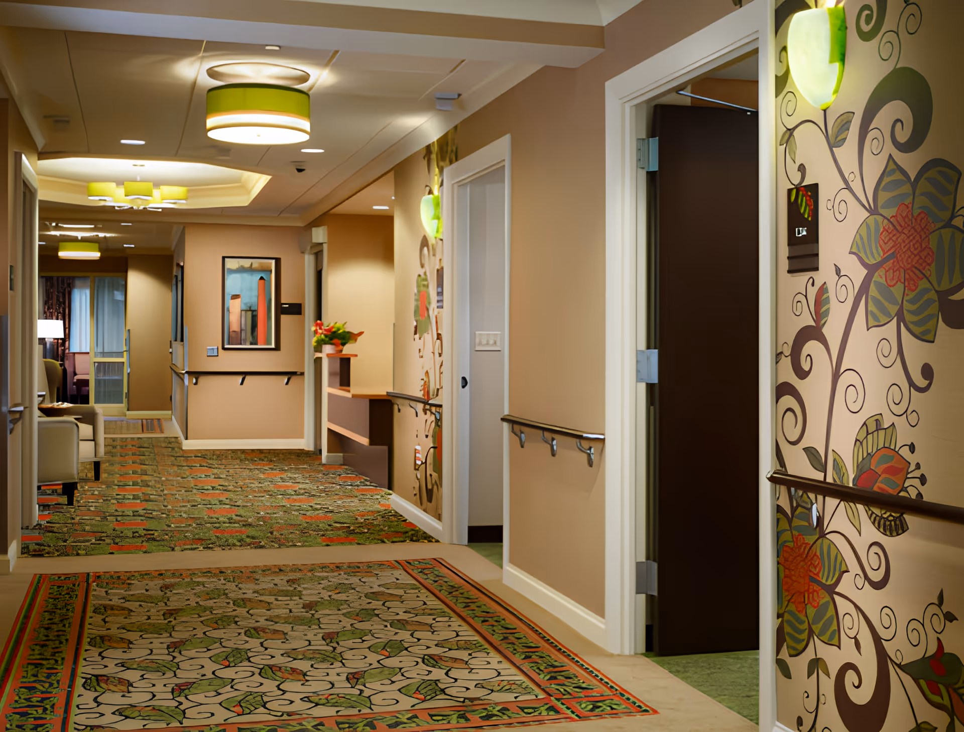 A well-lit hallway in a senior living facility with patterned carpet and beige walls. The hallway features handrails on both sides, decorative wall art with floral designs, ceiling light fixtures with green accents, and a framed picture on the far wall. There is a small seating area visible at the end of the hallway.