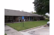 Single-story senior living facility front with green lawn, walkway, and a parked car nearby.