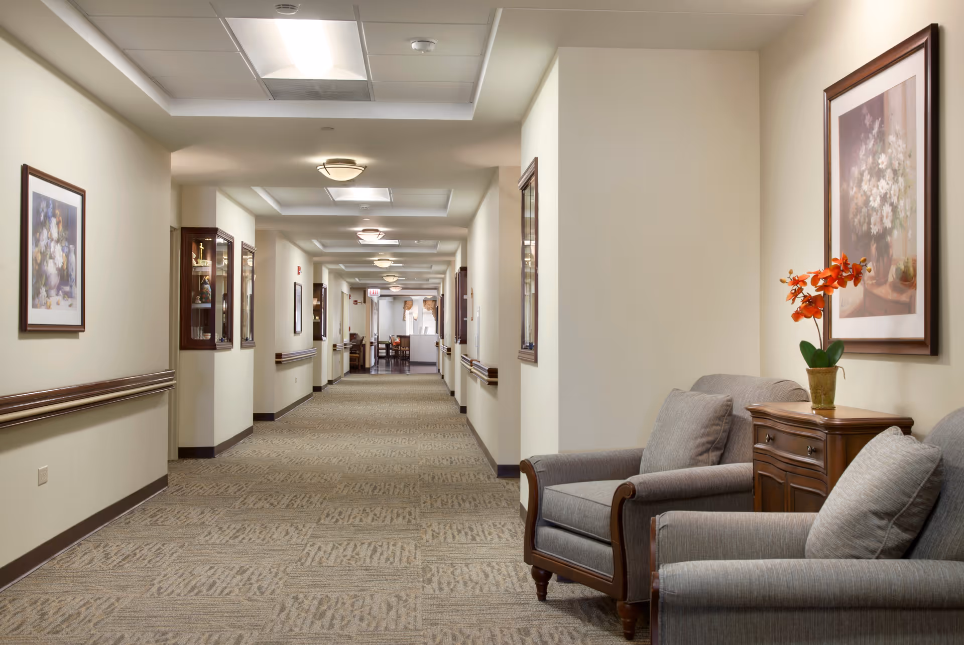 A long, well-lit hallway in a senior living facility with beige walls and carpeted floor. On the right side, there are two gray upholstered armchairs with a wooden side table between them, holding a vase with orange flowers. The walls are decorated with framed floral paintings and display cabinets. The hallway extends into a dining or common area visible in the distance.