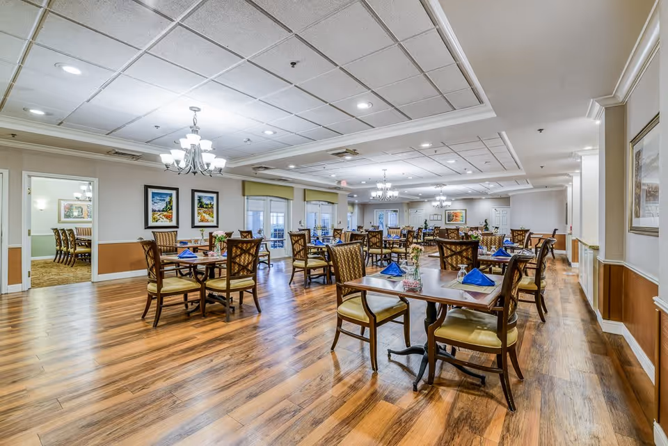 A spacious dining room with multiple wooden tables and chairs arranged neatly. Each table is set with blue napkins and small floral centerpieces. The room features wooden flooring, chandeliers hanging from a white ceiling with recessed lighting, and framed artwork on the walls. Large windows and glass doors allow natural light to enter the room.
