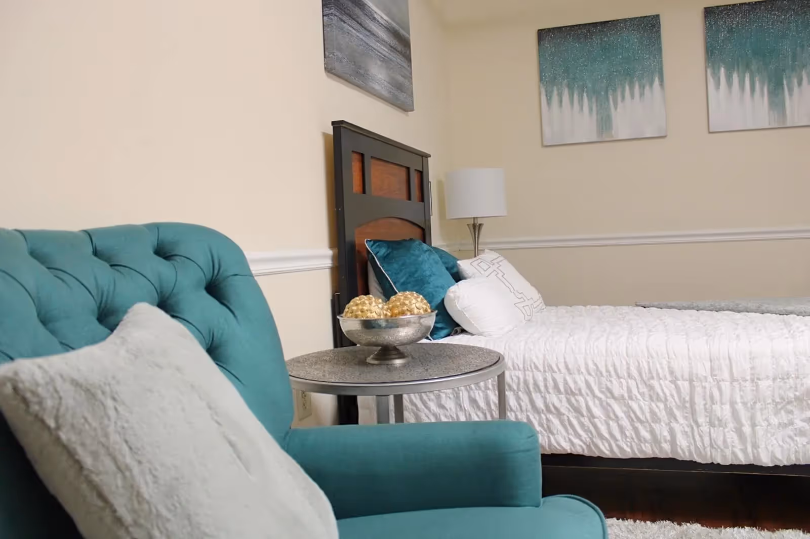 A cozy bedroom featuring a teal upholstered armchair with a light gray pillow, a round side table with decorative golden spheres in a silver bowl, a bed with white textured bedding and teal and white pillows, a wooden headboard, a floor lamp, and abstract artwork on the beige walls.