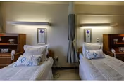 A room with two single beds placed side by side, each with white bedding and decorative pillows. Each bed has a wooden headboard with a small shelf holding a telephone and other items. Above each bed is a wall-mounted light fixture and a framed picture. A curtain hangs between the beds, and the walls are painted a light color.