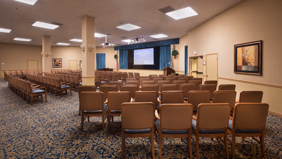 A large conference or meeting room with rows of brown chairs facing a stage with a podium and a projection screen displaying a presentation titled 'Busting Hurricane Myths'. The room has beige walls, blue patterned carpet, and ceiling lights. There are framed artworks on the walls and an American flag next to the stage.