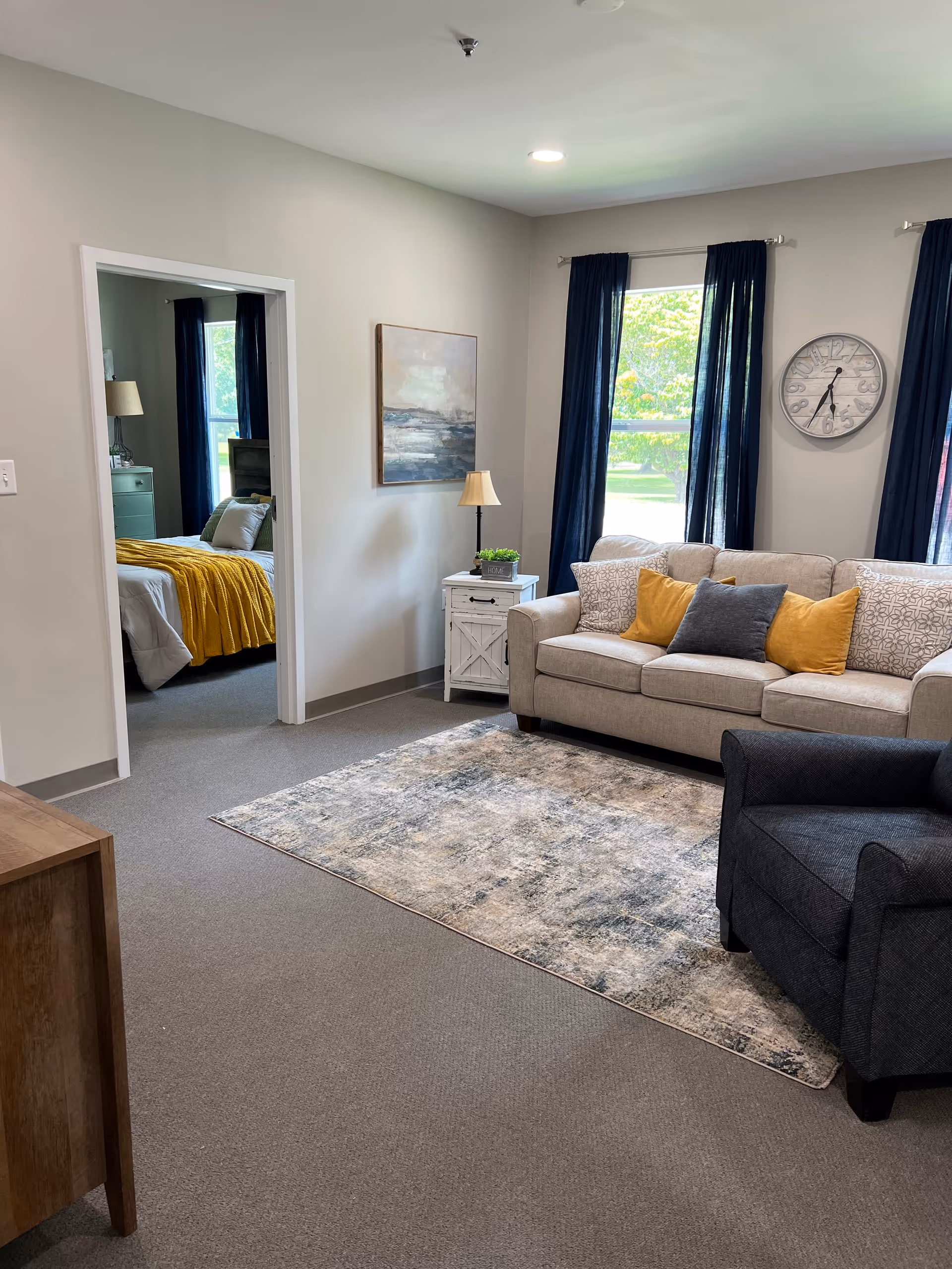 Living room with a beige sofa, dark armchair, area rug and side table, with a doorway showing a bedroom with a yellow throw.