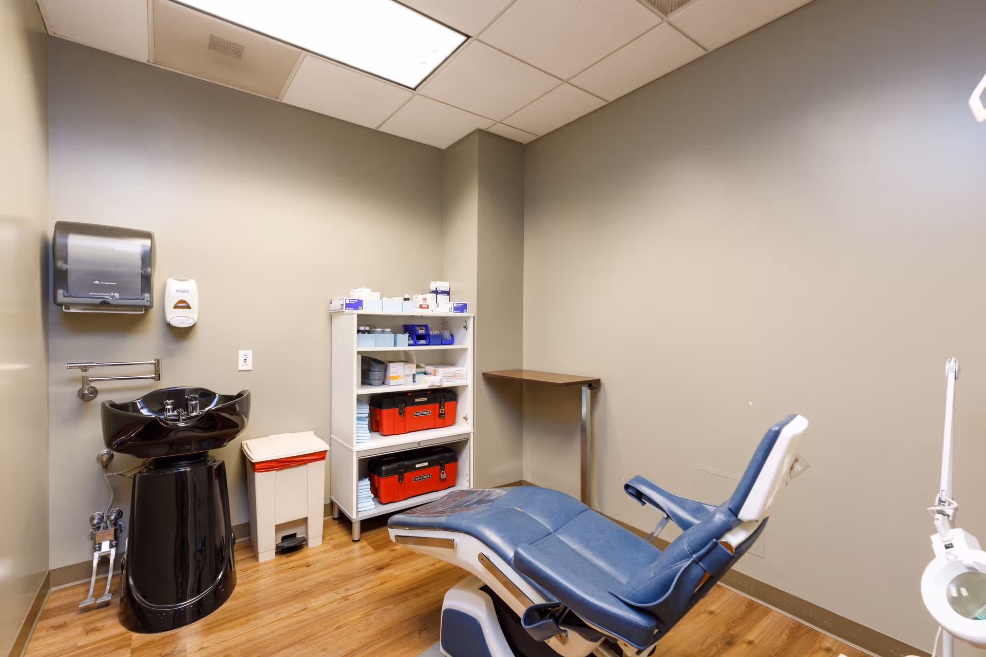 A small medical or dental examination room with a blue reclining chair, a black sink, a white shelving unit stocked with medical supplies, a trash bin, and a wall-mounted paper towel dispenser and soap dispenser. The room has beige walls and a wooden floor.