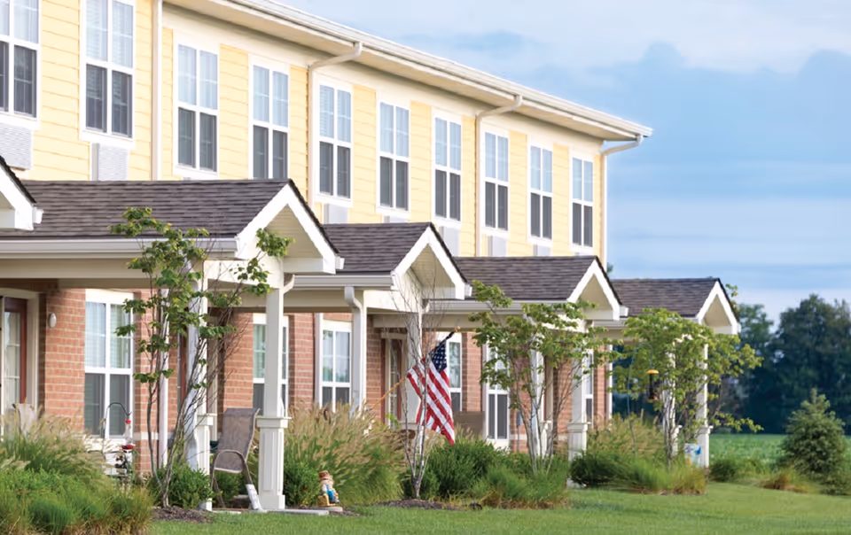 Exterior view of a senior living facility with a row of attached units featuring covered porches, small trees, and an American flag displayed on one porch. The building has yellow siding on the upper level and brick on the lower level, with multiple windows and a well-maintained lawn in front.