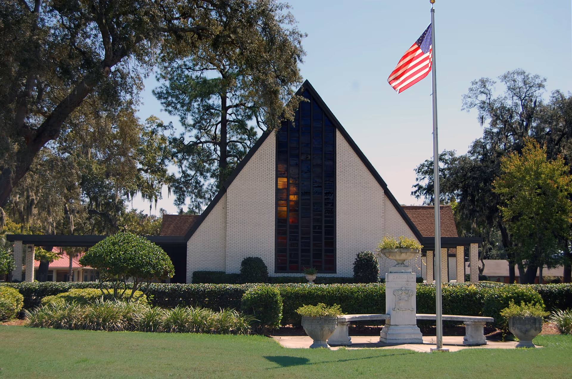 A triangular-shaped building with a tall, narrow window in the center, surrounded by well-maintained bushes and trees. In front of the building is a flagpole with an American flag and a stone monument with benches around it. The sky is clear and blue.