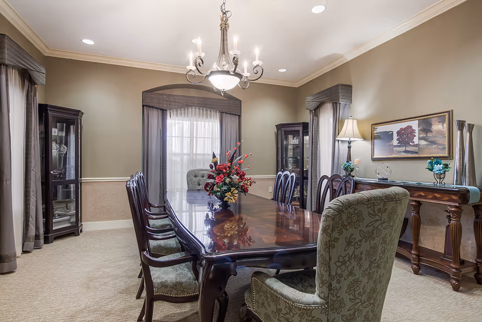 Elegant dining room with a long polished wooden table surrounded by upholstered chairs. A floral centerpiece decorates the table. The room features beige walls, carpeted floor, two glass display cabinets, a chandelier, a side table with a lamp, decorative vases, and framed artwork on the wall. Large windows with sheer curtains allow natural light to enter.