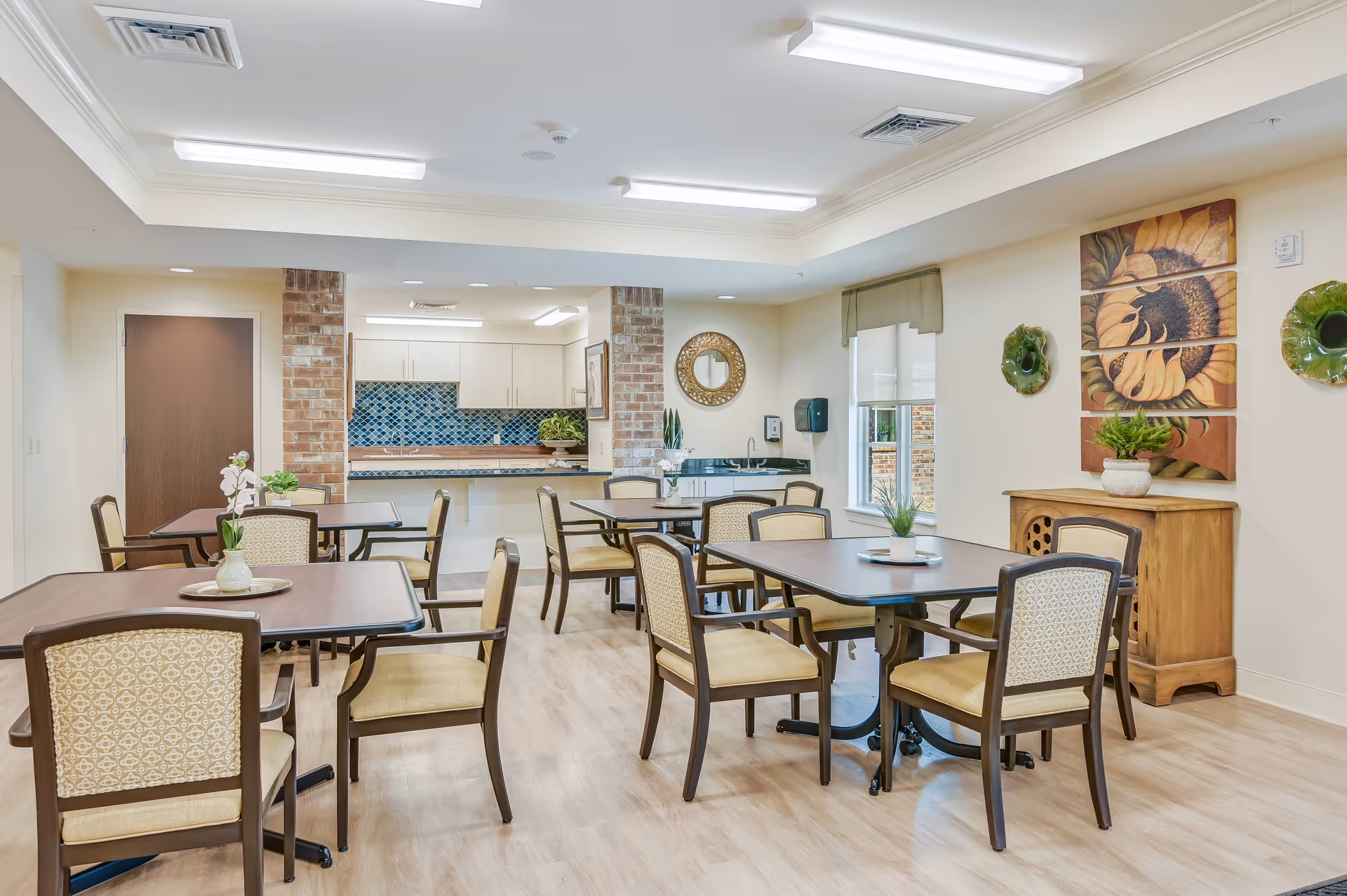 A bright dining room in a senior living facility with several wooden tables and cushioned chairs arranged neatly. The room features light-colored walls, a wooden floor, decorative wall art including a sunflower painting and green ceramic pieces, and a small wooden cabinet. In the background, there is a kitchen area with white cabinets and a blue tiled backsplash, separated by two brick columns.