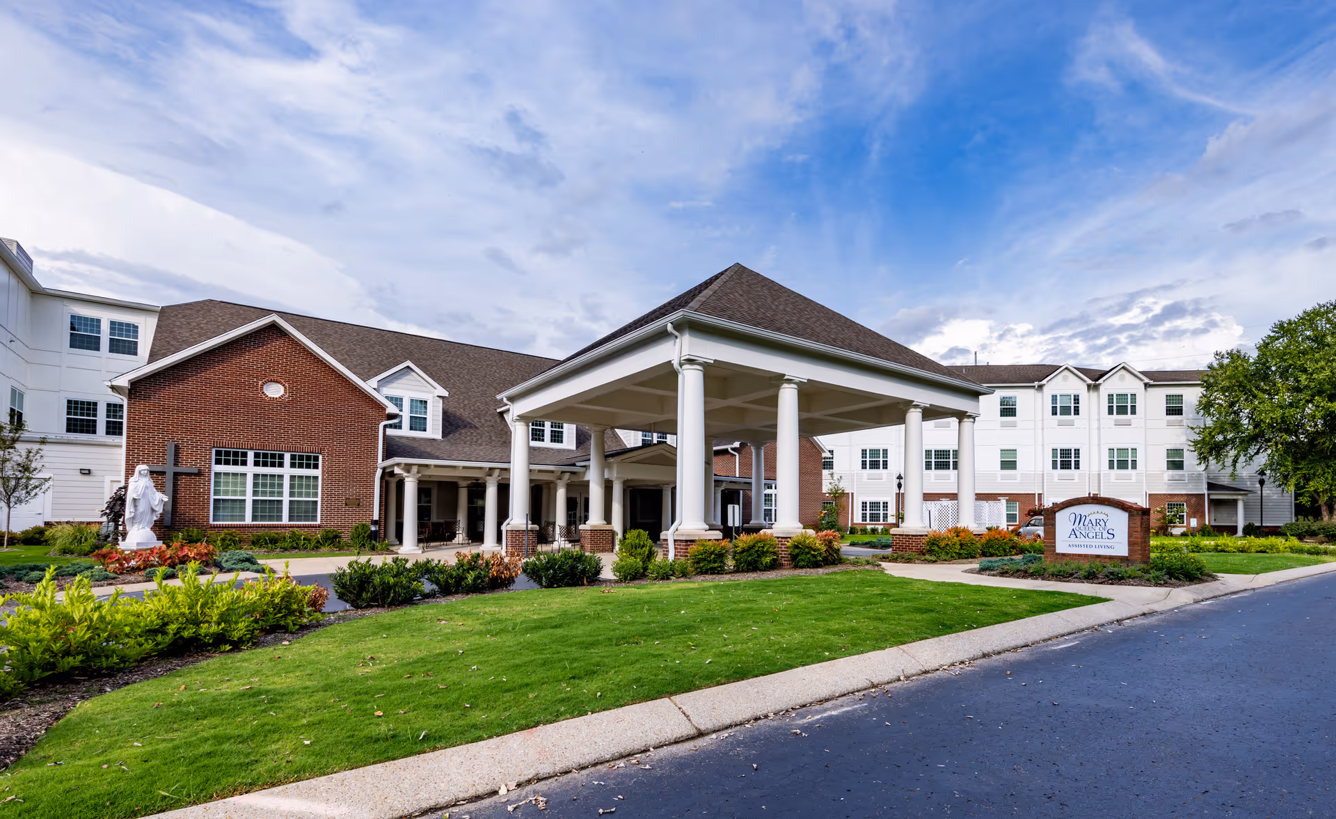 Exterior view of Mary Queen of Angels Assisted living facility showing a large covered entrance with white pillars, a well-maintained lawn, shrubs, and a statue near the building. The building has a combination of brick and white siding with multiple windows under a partly cloudy sky.