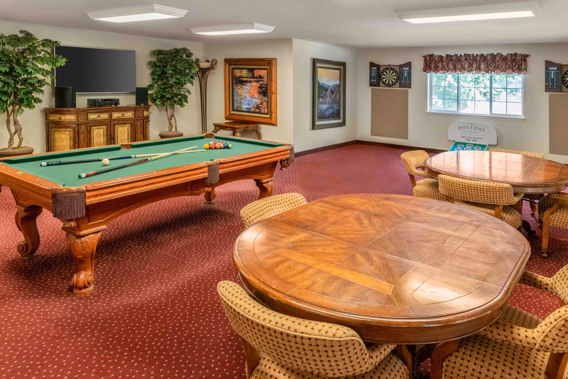 A recreational room with a pool table in the center, two wooden tables with cushioned chairs around them, a TV on a wooden cabinet, two artificial plants, framed paintings on the walls, a dartboard, and a window with a valance letting in natural light.