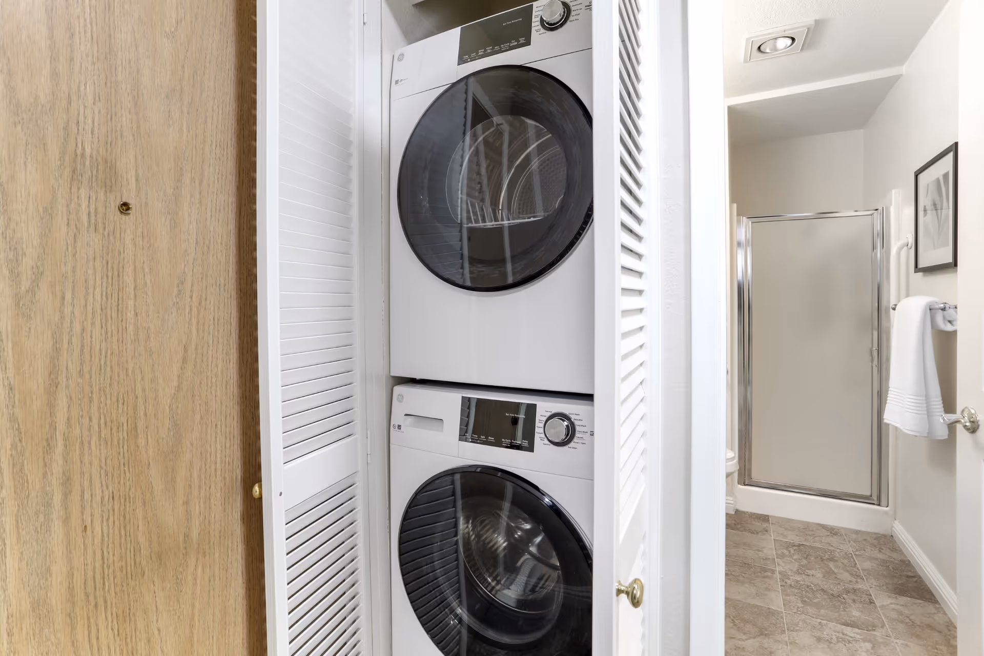 A stacked washer and dryer unit inside a white closet with louvered doors. To the right, there is a bathroom with a glass shower door, a towel hanging on a rack, and a framed picture on the wall. The floor is tiled in a light brown color.