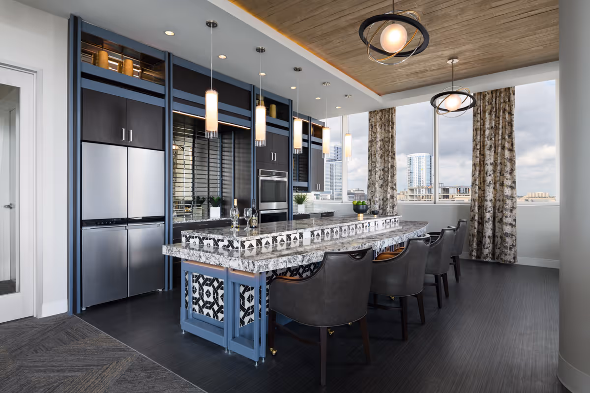 Modern kitchen area with a large granite island countertop featuring black and white patterned tiles on the sides. The island is surrounded by six dark leather chairs. The kitchen has dark cabinetry with stainless steel appliances including a refrigerator and built-in oven. Pendant lights hang above the island, and large windows with patterned curtains provide a cityscape view.