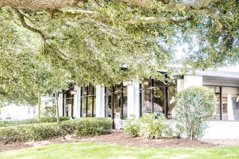 Exterior view of a senior living facility facade with large windows, manicured shrubs, and overhanging trees.