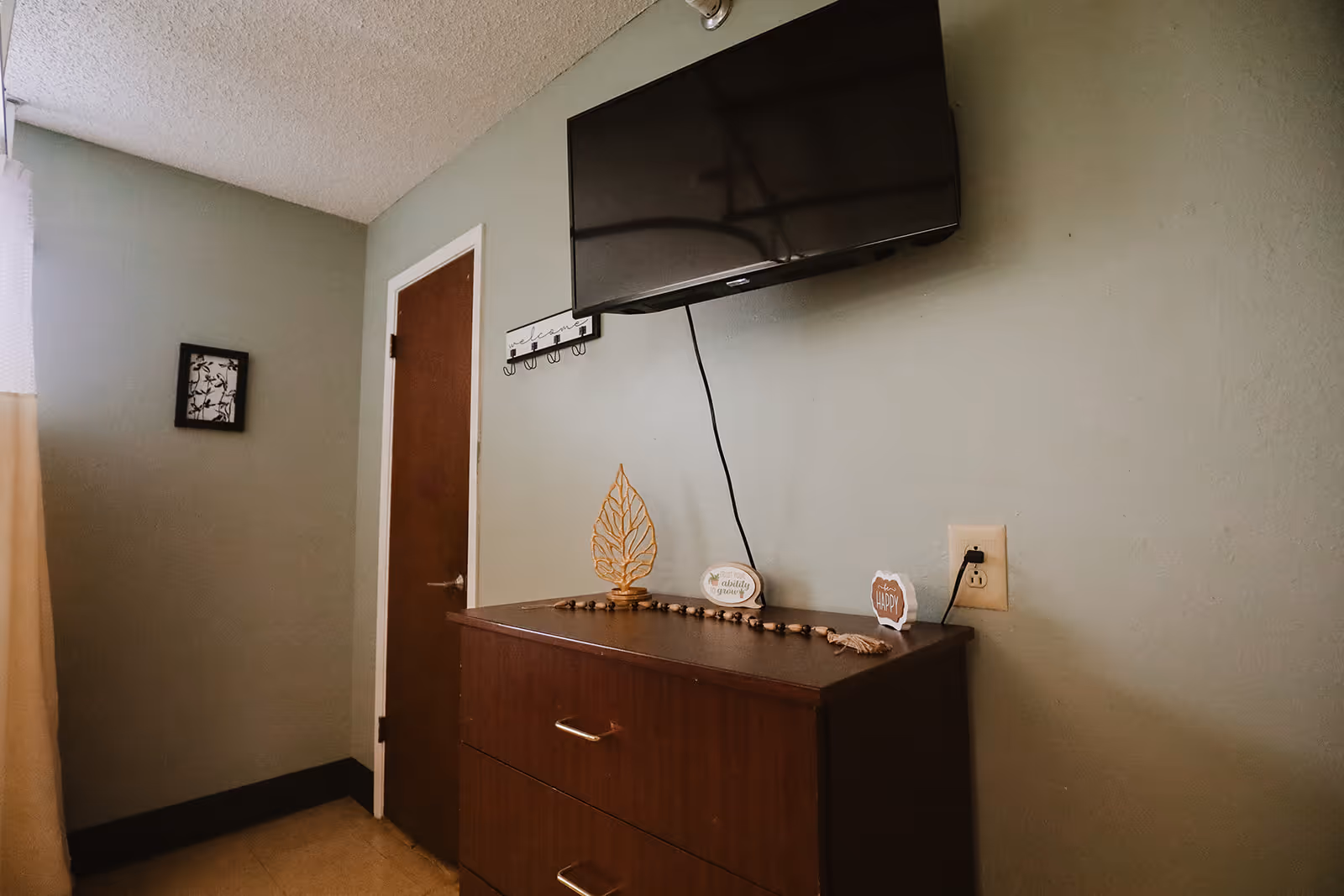 A small room with a mounted flat-screen TV above a wooden dresser. On the dresser are decorative items including a leaf sculpture, a beaded garland, and small signs with positive messages. There is a closed wooden door and a small framed picture on the wall. The walls are painted light green.