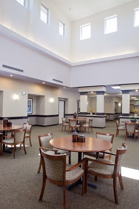 A spacious dining area with round wooden tables and cushioned chairs arranged neatly. The room has high ceilings with multiple small windows near the top, allowing natural light to brighten the space. The walls are painted in neutral tones with dark trim, and there are decorative columns and wall sconces providing additional lighting.