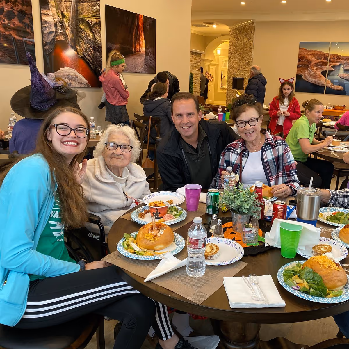 A group of people, including elderly and younger adults, sitting around a round table enjoying a meal together in a communal dining area. The table is set with plates of food, drinks, and a small decorative plant. The background shows other people seated and standing, with wall art and warm lighting creating a cozy atmosphere.