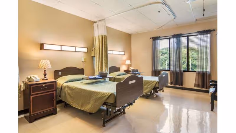 Shared patient bedroom with two adjustable beds, bedside tables and lamps, a privacy curtain, and a window with blinds.