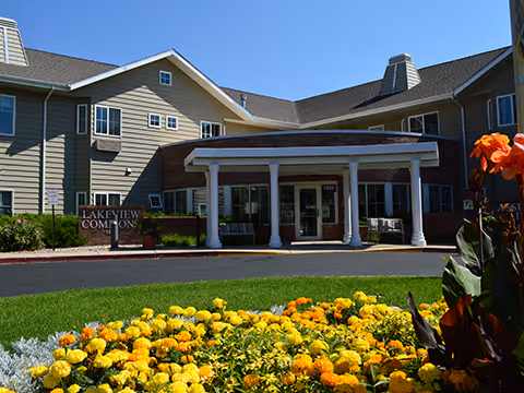 Front exterior view of Lakeview Commons building with a covered entrance, surrounded by a well-maintained garden featuring yellow and orange flowers in the foreground.