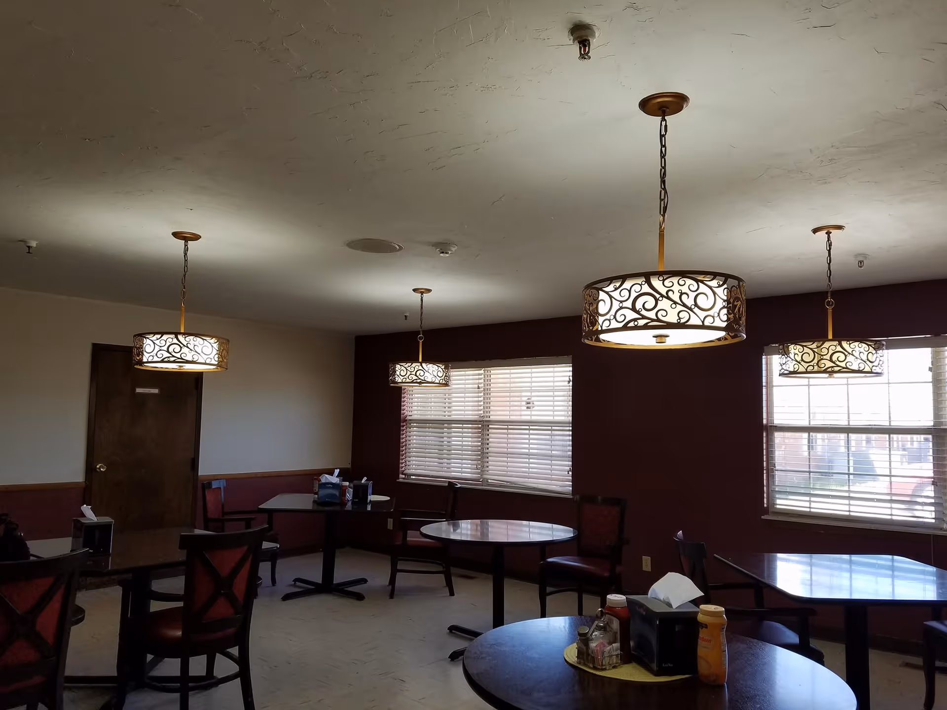 Interior view of a dining room with multiple round and square tables and chairs. The room has three decorative hanging light fixtures with ornate designs, windows with blinds letting in natural light, and a door on the left side.