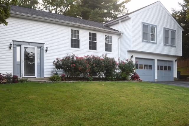 Exterior view of a white two-story house with gray shutters and a double garage. There is a green lawn in front and a row of flowering bushes along the house.
