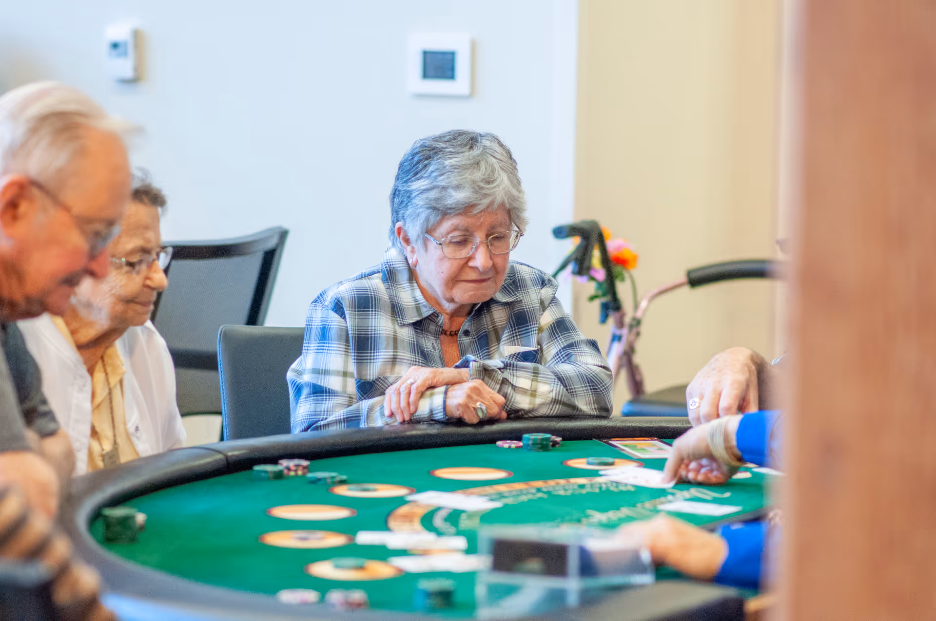 A group of elderly people sitting around a green felt gaming table, engaged in a card game. One woman in a plaid shirt is focused on the game, while others watch and participate. A walker with flowers is visible in the background.