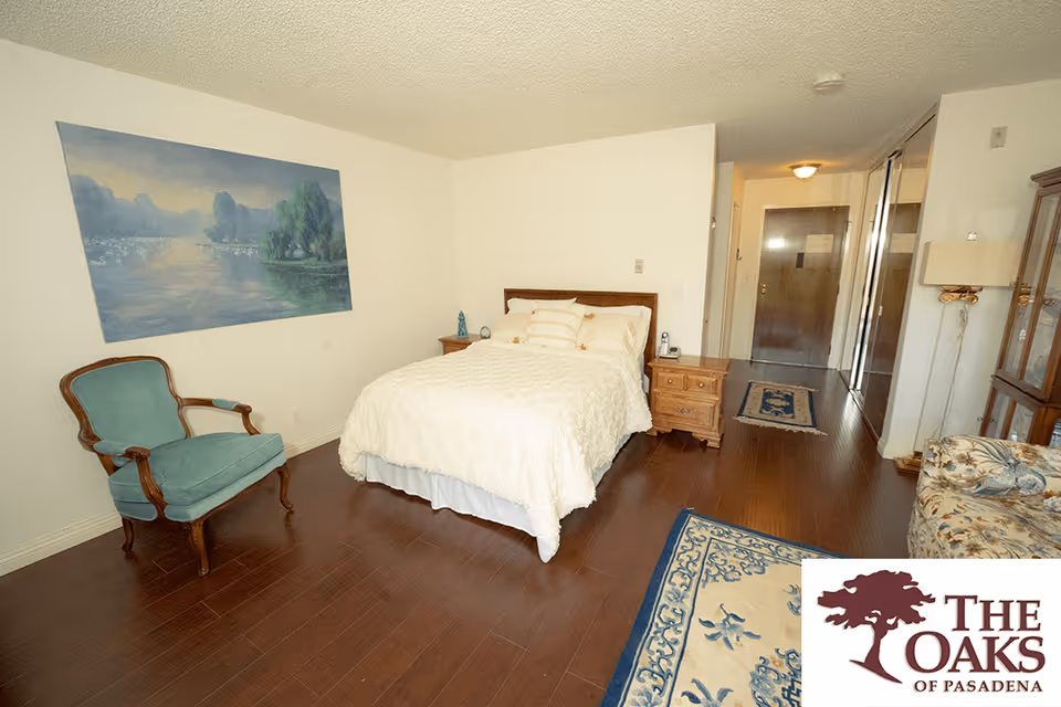 A bedroom in a senior living facility with a bed covered in a white quilt, a wooden nightstand with a lamp and clock, a blue upholstered armchair, a floral patterned armchair, a large painting of a lake scene on the wall, hardwood floors, and a hallway leading to a door. A rug with a blue and beige pattern is partially visible on the floor.