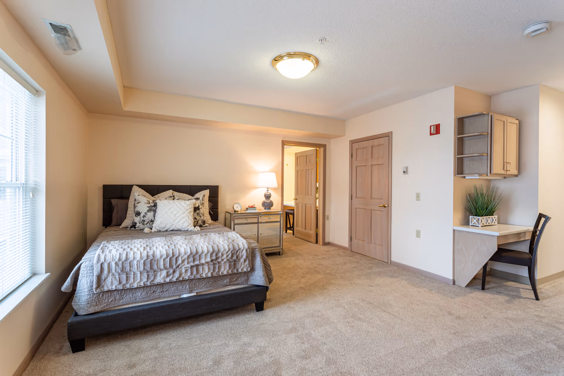 A spacious bedroom with a large bed adorned with multiple pillows and a textured blanket. Next to the bed is a mirrored nightstand with a lamp and a clock. The room has beige walls and carpeted floors. There is a window with blinds on the left side letting in natural light. On the right side, there is a small built-in desk with a chair and a potted plant. Two wooden doors are visible, one open leading to another room and one closed.