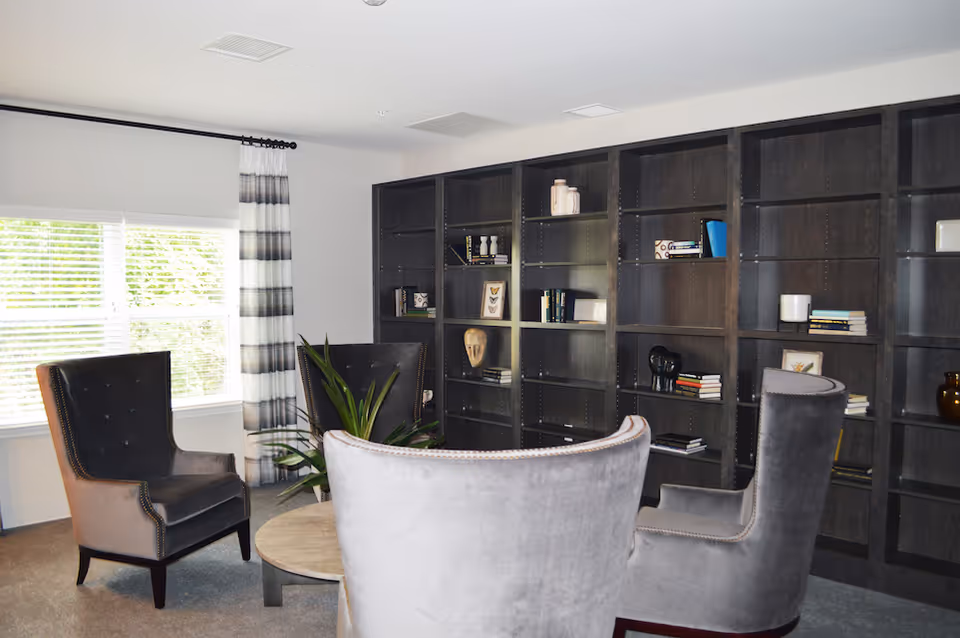 Seating area with gray upholstered armchairs around a low round table in front of large built-in dark wood bookshelves.
